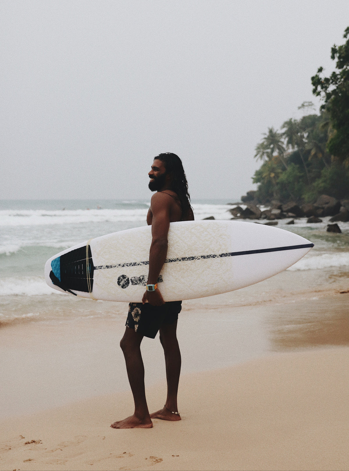 Sahan standing at the shoreline with a white surfboard, looking toward small breaking waves