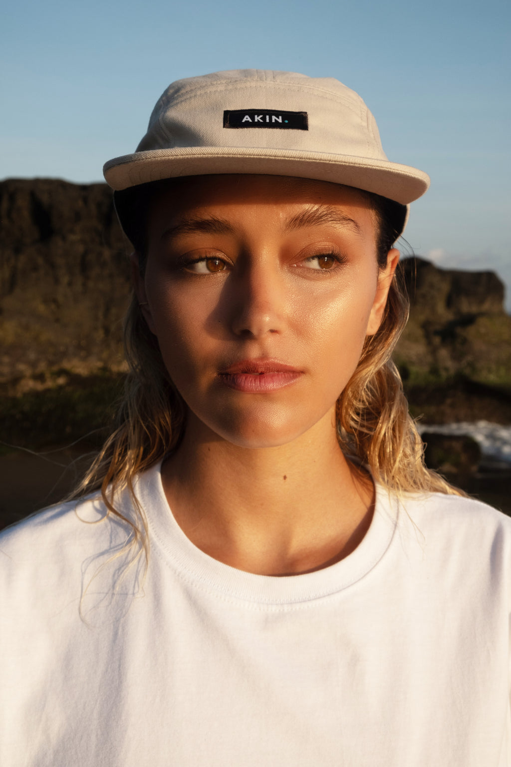 Model wearing beige Akin Five Panel Cap outdoors, facing camera against rocky natural backdrop