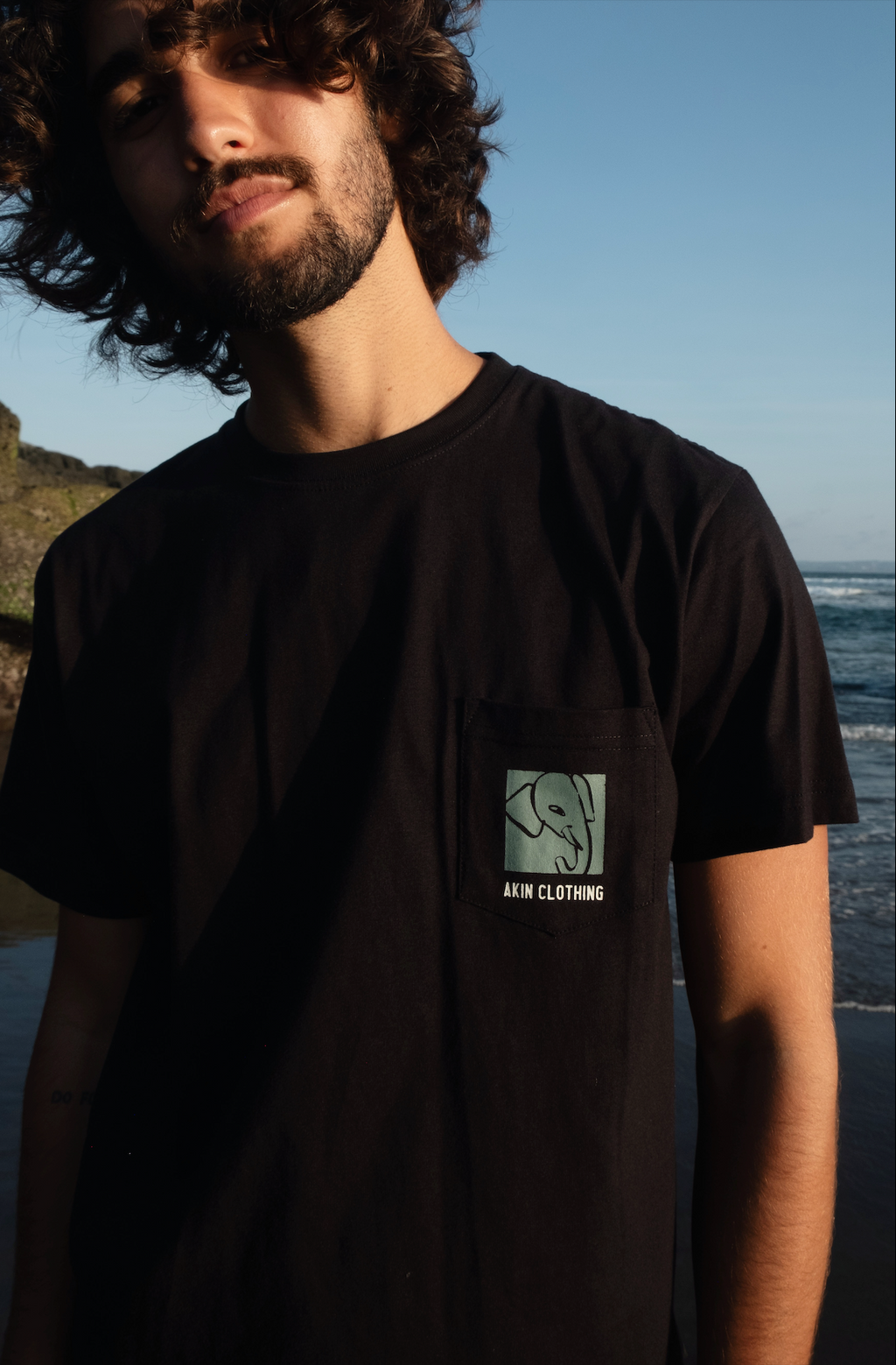 Man wearing Sahan Pocket T-Shirt black, elephant illustration and Akin Clothing on chest pocket, coastal backdrop