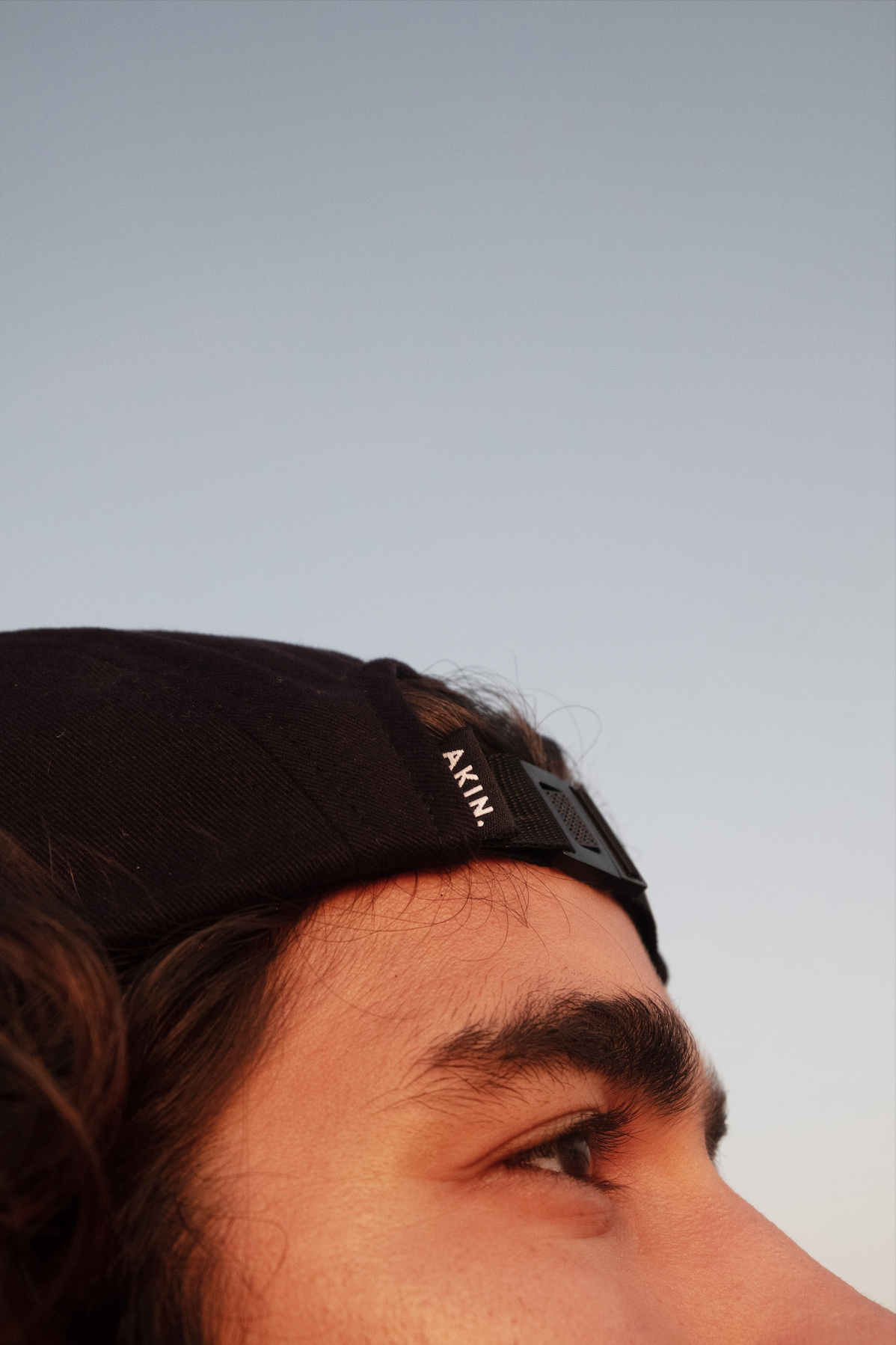 Model wearing black Akin Five Panel Cap, side profile close-up against pale blue sky