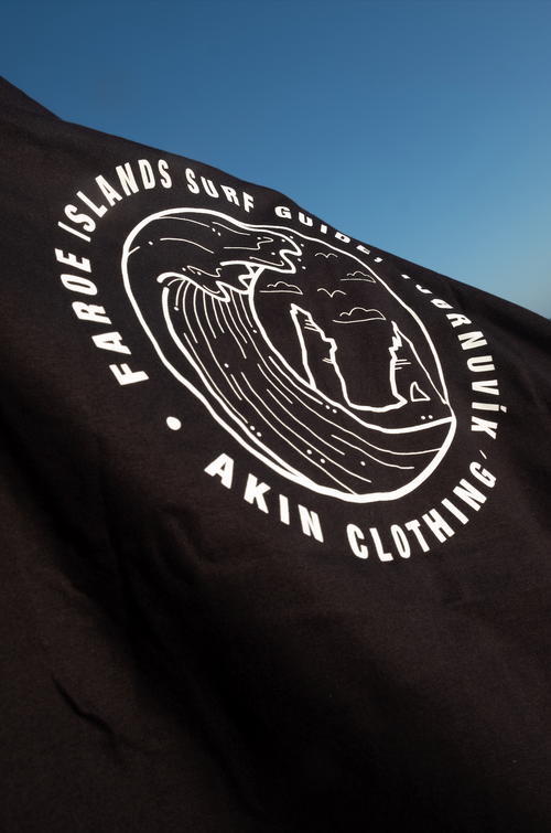 Close-up of circular Faroe Islands Surf Guide graphic on black Akin t-shirt against blue sky