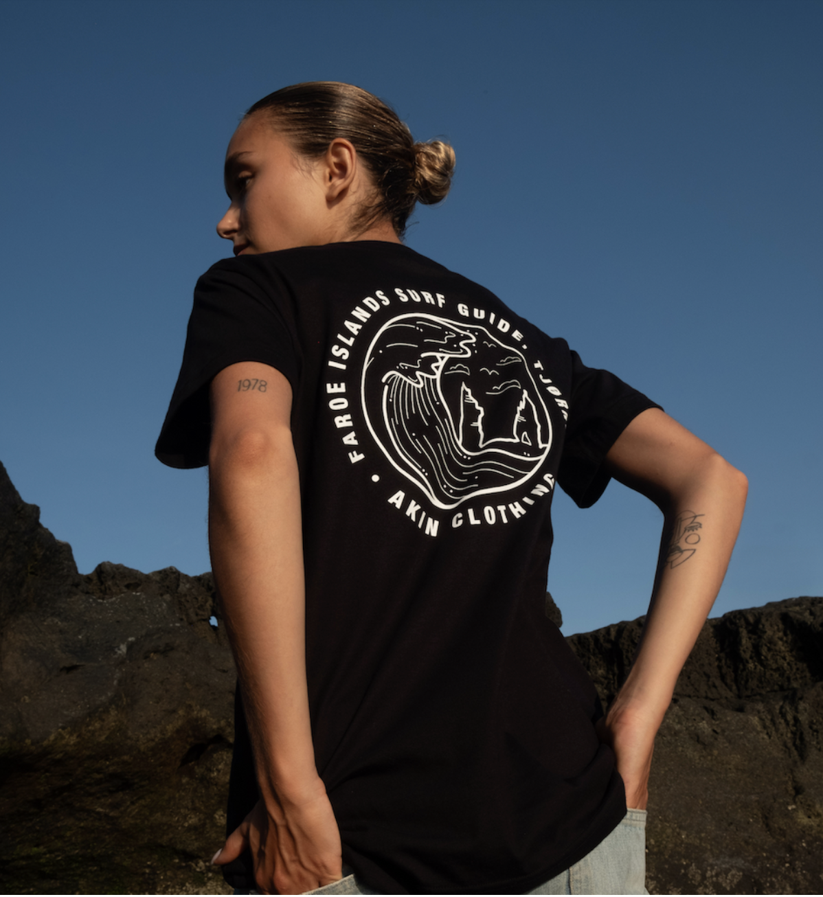 Model wearing black Faroe Islands Surf Guide x Akin t-shirt, back view, rocky landscape