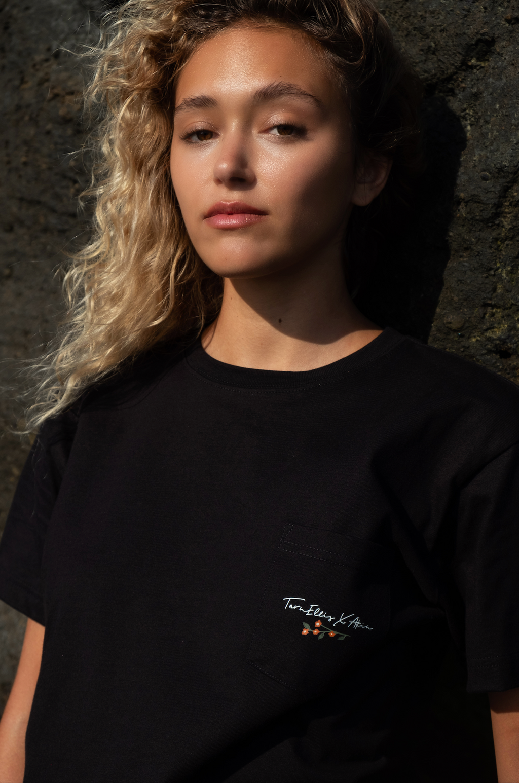 Woman wearing Tarn Ellis x Akin black pocket t-shirt, signature script and floral chest graphic, stone backdrop